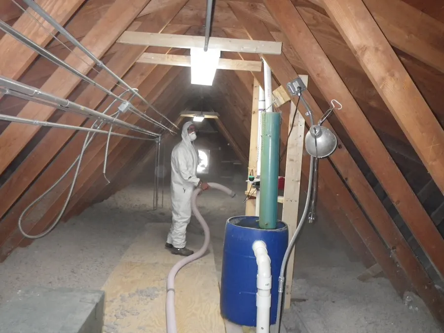 Unfinished attic with wooden rafters, conduit along the beams and a worker in a protective suit blowing loose insulation across the floor