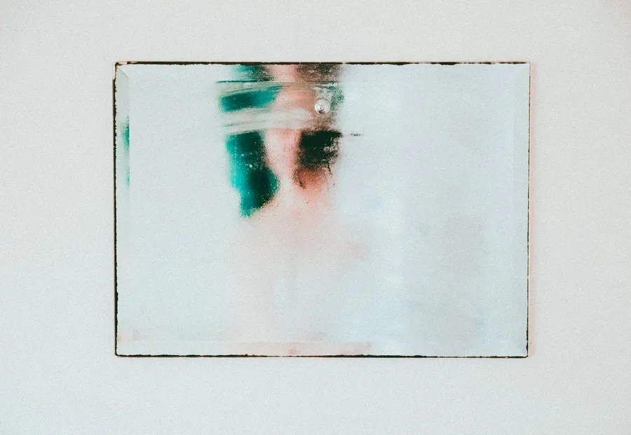Bathroom mirror on a white wall covered in condensation, with a blurred reflection behind the fog