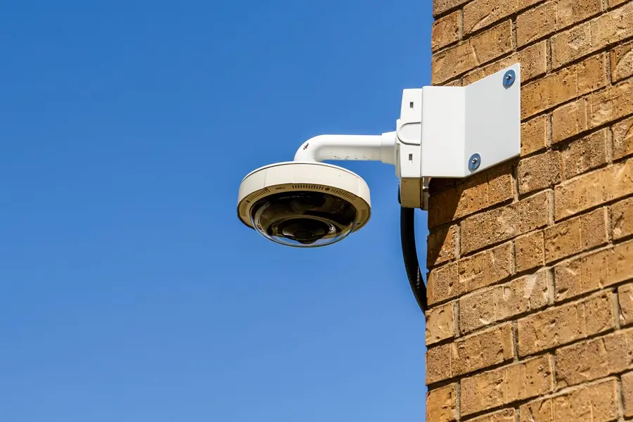 White dome security camera on a wall bracket fixed to a tan brick wall, its cable running down the bracket into the wall