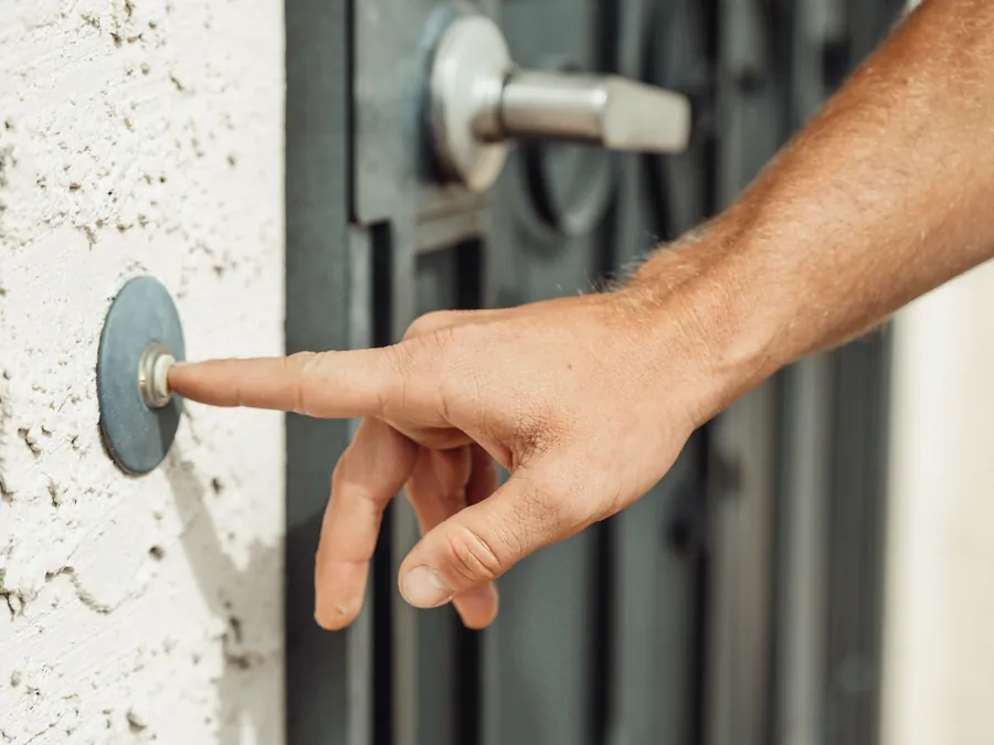 A finger pressing a round push-button doorbell on a white stucco wall beside a dark metal front door
