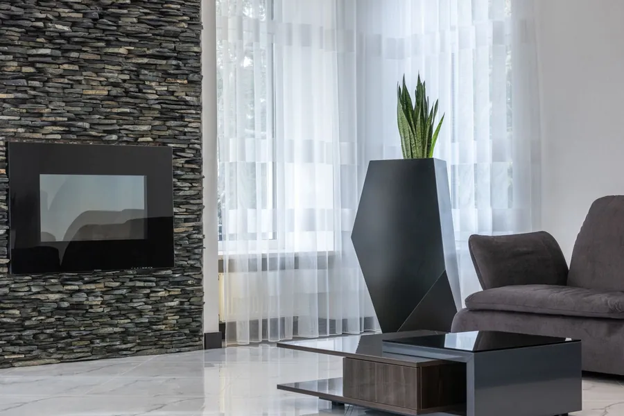 Black wall-mounted electric fireplace set into a stacked-stone wall beside tall windows and a grey sofa