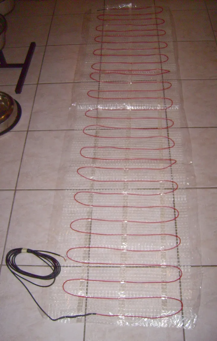 Electric floor heating mat with a red heating wire looped across clear mesh, laid out on a white tile floor with its black power lead coiled at one end