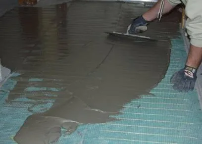 Gloved installer spreading gray thinset with a trowel over blue mesh floor heating mats covering a subfloor