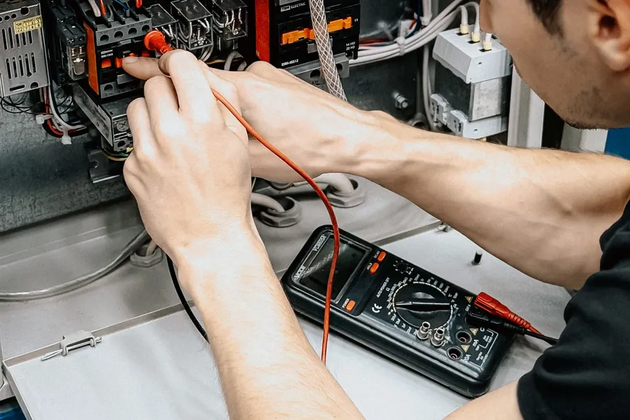 Electrician's hands holding a red test lead against the terminals of an open control panel, a digital multimeter resting on the shelf below