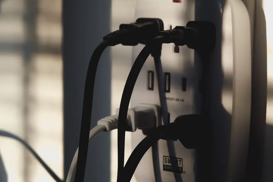 Wall outlet tap with several black and white plugs and cords hanging from it, lit by low sun through blinds