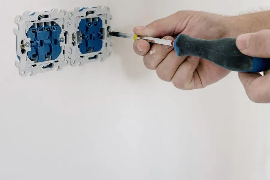 Electrician's hands tightening a terminal screw on a pair of open receptacle boxes set in a white wall