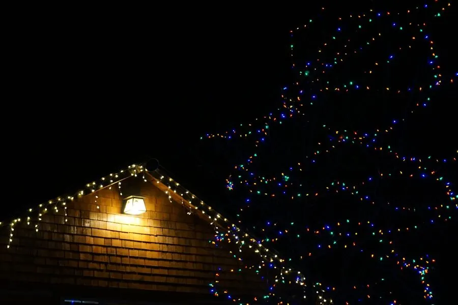 Warm white icicle lights hung along the eaves of a shingled gable at night, with a lit tree behind the house