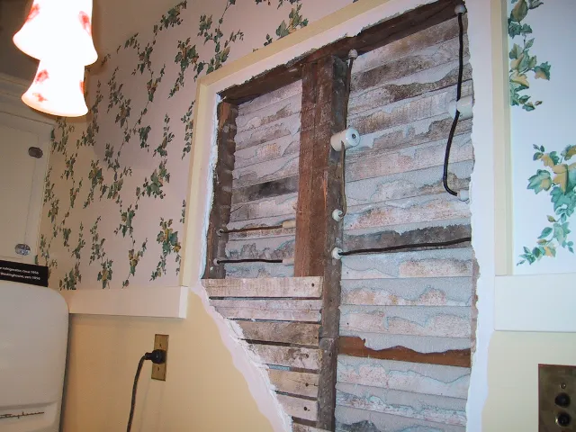 Opened plaster wall showing old cloth-covered wires on white porcelain knobs and tubes along wooden studs
