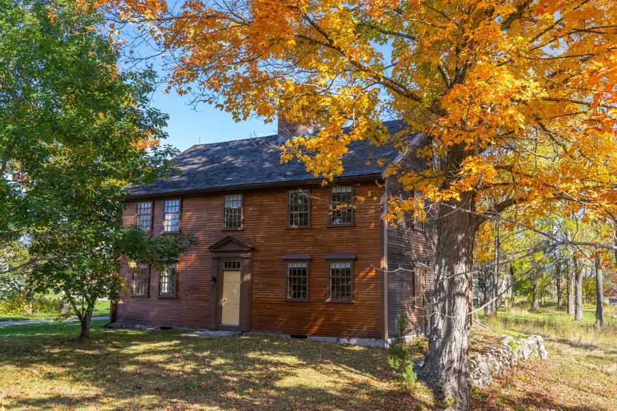 Two-story colonial-era house with brown clapboard siding under a large maple tree in orange fall leaves