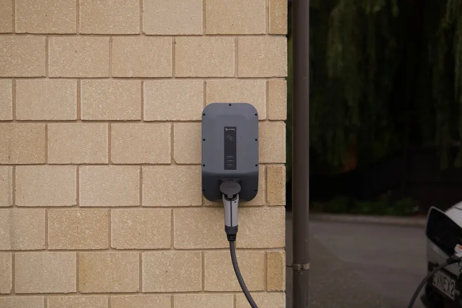 Gray wall-mounted electric vehicle charger with its cable plugged in, on a pale brick wall beside a parked car