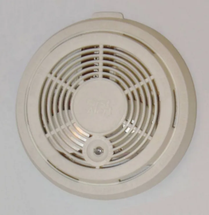 Older round smoke detector on a ceiling, its plastic cover yellowed with age