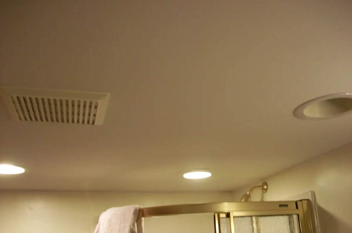 Bathroom ceiling with a white exhaust fan grille, two recessed lights and the top of a glass shower enclosure