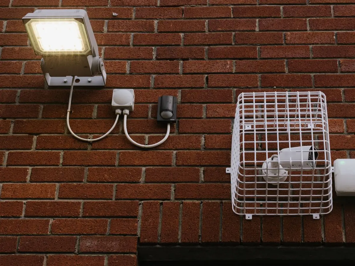 Brick wall with an LED floodlight and motion sensor wired through a white junction box, next to a caged bullet security camera