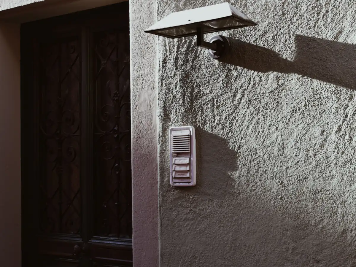 Stucco wall beside a dark wooden front door, with a doorbell panel at hand height and a wall lamp above it