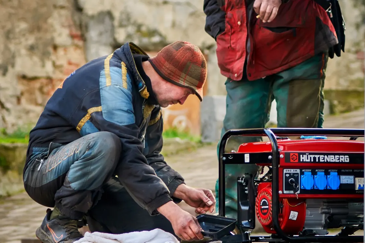 Technician crouched beside a red portable generator, working on it with a cloth laid out on the ground while a second worker stands by
