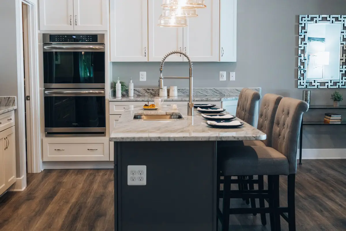 Kitchen with white cabinets, a stone-topped island with a sink and an outlet on the side of the island, and two built-in wall ovens