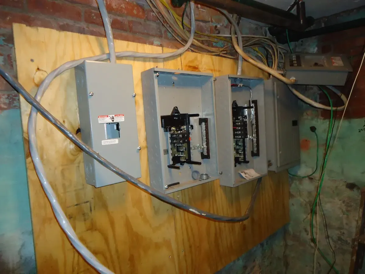 New breaker panels being installed on a plywood board in an old brick basement, with older wiring overhead