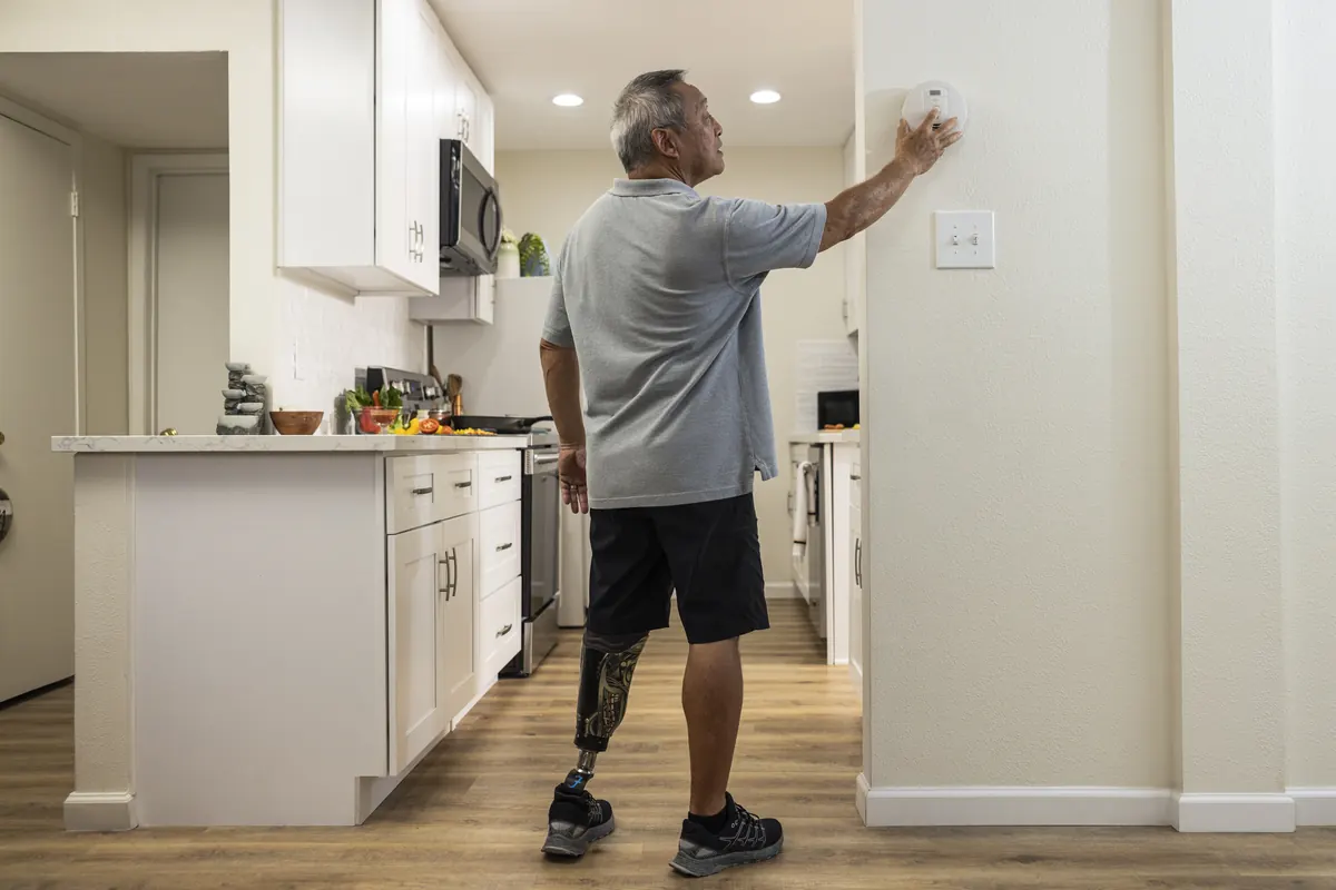 Man pressing the test button on a round smoke alarm mounted on a wall beside a kitchen